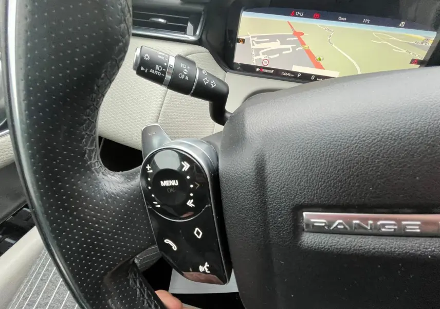 Gros plan sur le volant noir perforé du Range Rover Velar 2017, avec commandes intégrées et écran de navigation visible en arrière-plan.