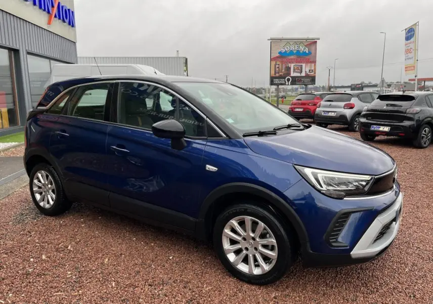Opel Crossland X bleu clair en vue 3/4 avant droit, avec toit noir et jantes argentées, stationné sur un sol en gravier.
