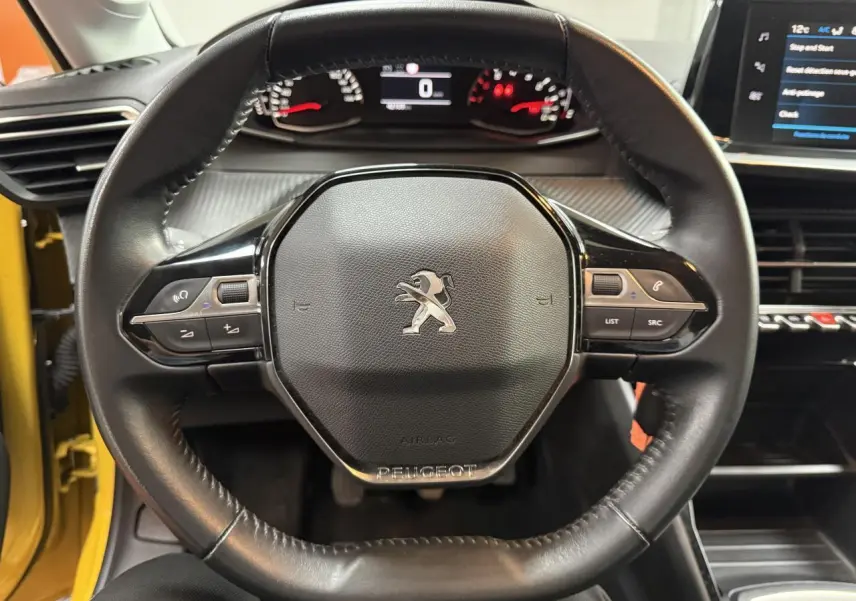 Vue centrée sur le volant cuir noir de la Peugeot 208 2020 avec tableau de bord et écran tactile visibles, portière jaune ouverte.
