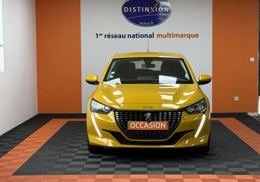 Vue frontale d'une Peugeot 208 jaune clair avec calandre noire et plaque "OCCASION" dans un showroom.