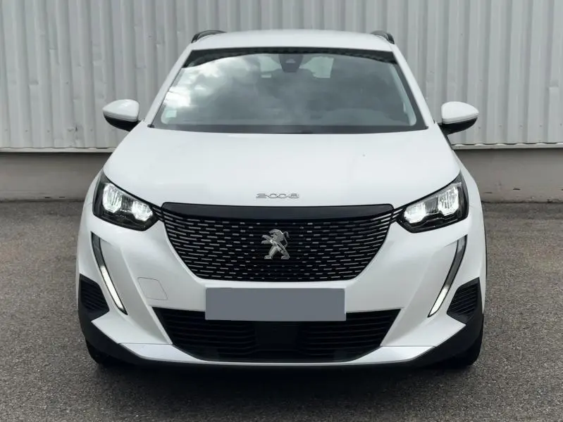 Vue frontale d'un Peugeot 2008 blanc banquise avec calandre noire et feux LED allumés sur fond urbain.
