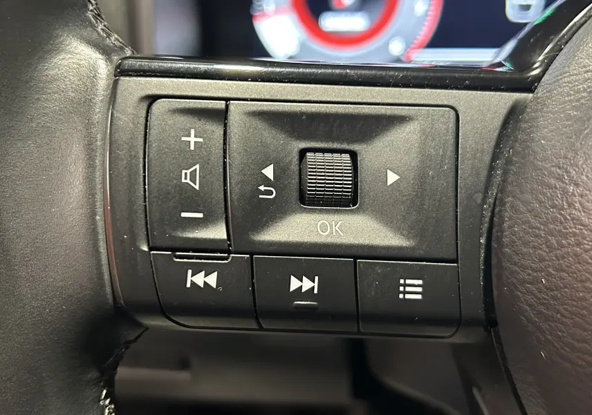 Gros plan sur les commandes noires du volant côté gauche du Nissan Qashqai 2023, avec affichage numérique flou en arrière-plan.