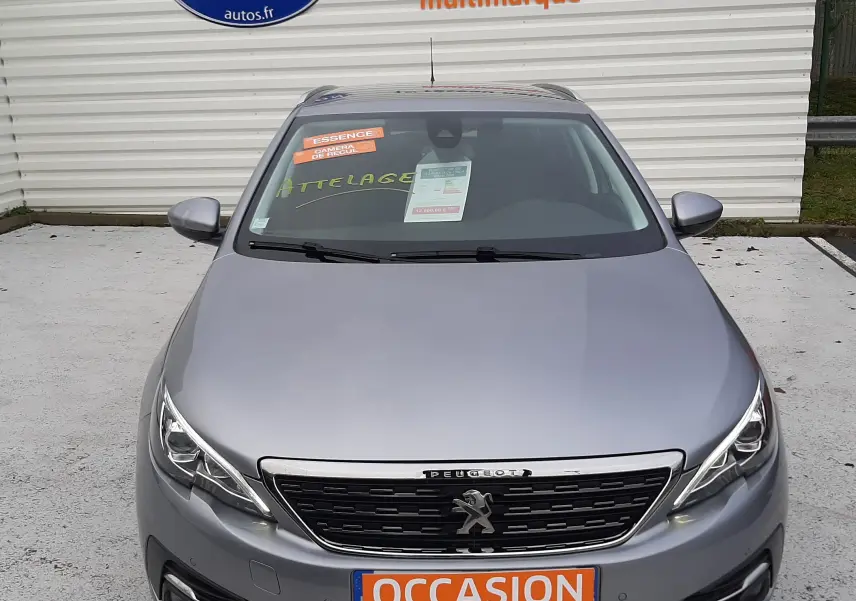 Vue avant d'une Peugeot 308 SW gris Artense avec plaque "OCCASION" et autocollants sur pare-brise dans un parking.