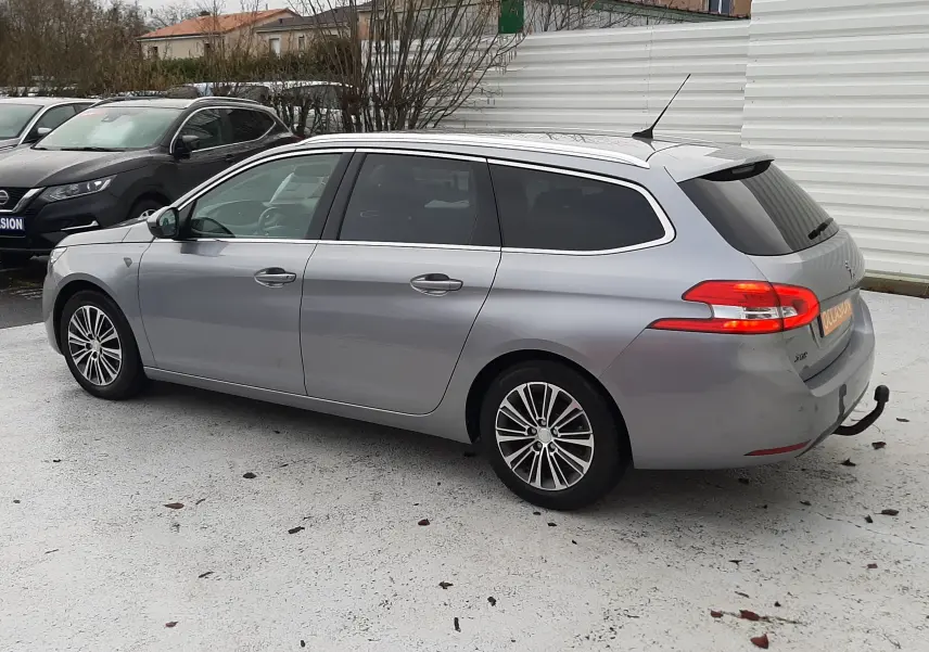 Peugeot 308 SW gris Artense vue 3/4 arrière côté gauche, break avec feux arrière allumés et attelage visible.