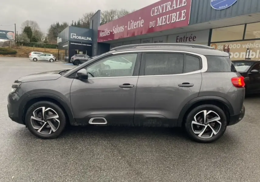 Vue de profil côté gauche d'un Citroën C5 Aircross gris platinium avec jantes alliage 18 pouces, garé devant un magasin.