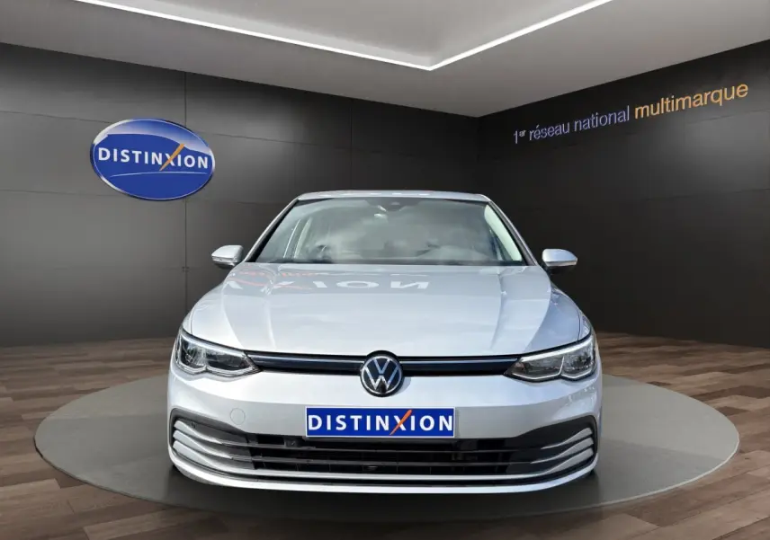 Vue frontale d'une Volkswagen Golf gris clair 2022 avec logo VW et plaque Distinxion visible en showroom.