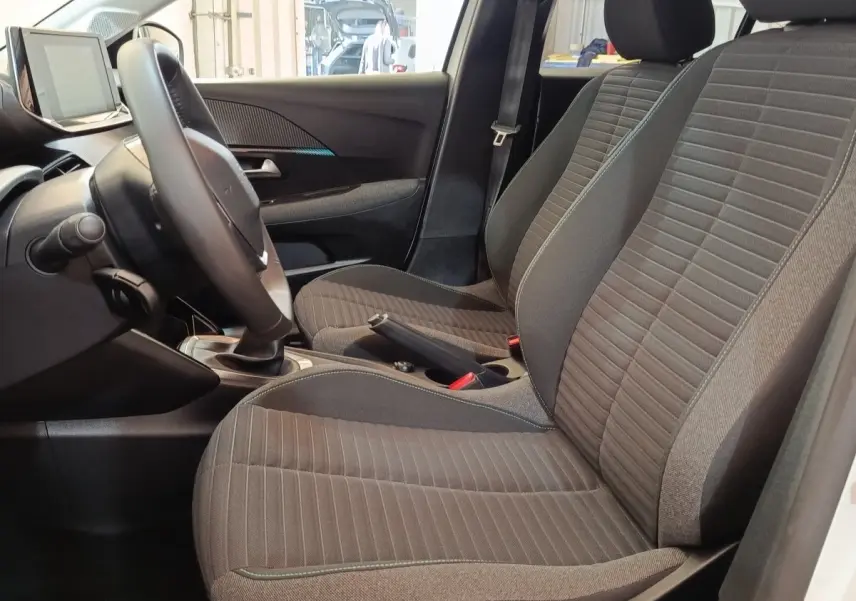 Vue intérieure côté conducteur de la Peugeot 208 blanche 2023, sièges tissu gris et volant cuir multifonctions.