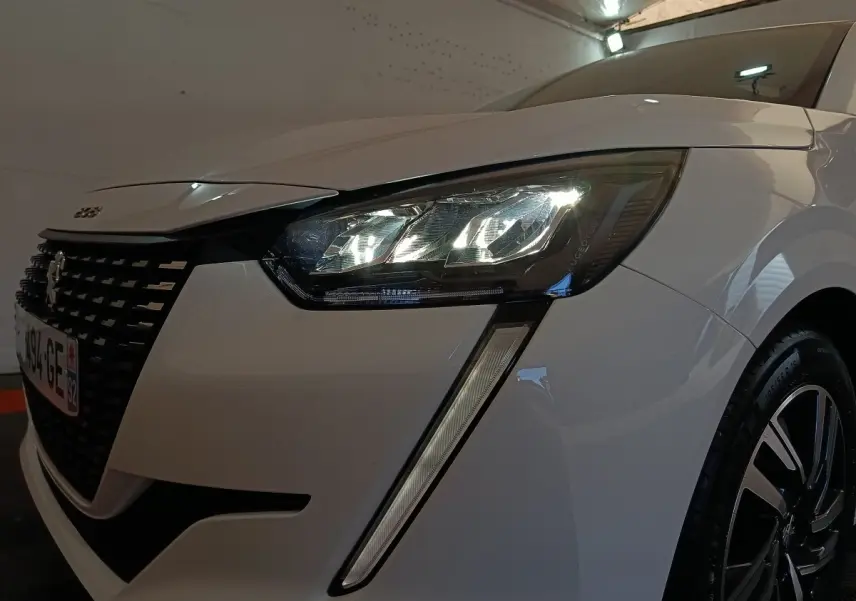 Gros plan sur l'avant droit d'une Peugeot 208 blanche 2023, mettant en valeur son phare LED et la calandre distinctive.