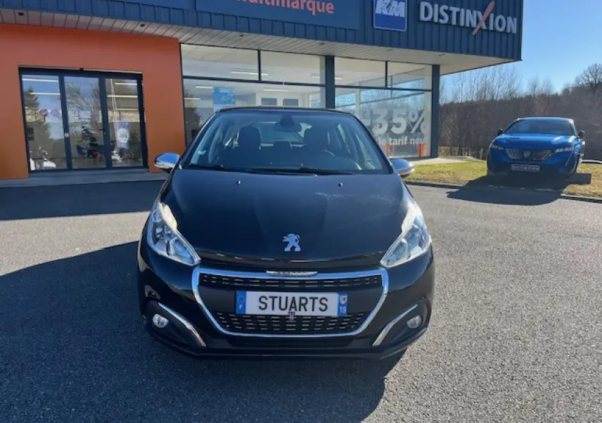 Vue avant d'une Peugeot 208 noire garée devant un bâtiment commercial avec calandre chromée et phares allumés.