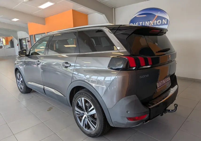 Peugeot 5008 gris vue 3/4 arrière droit en intérieur, avec feux arrière à griffes rouges et attelage visible.