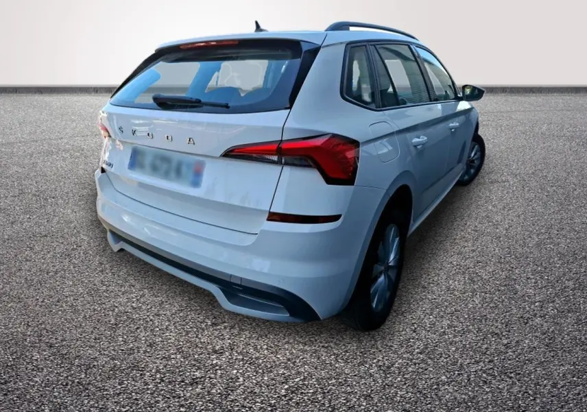 Vue 3/4 arrière droite d'un Skoda Kamiq blanc avec feux LED et barres de toit noires sur sol gravillonné.