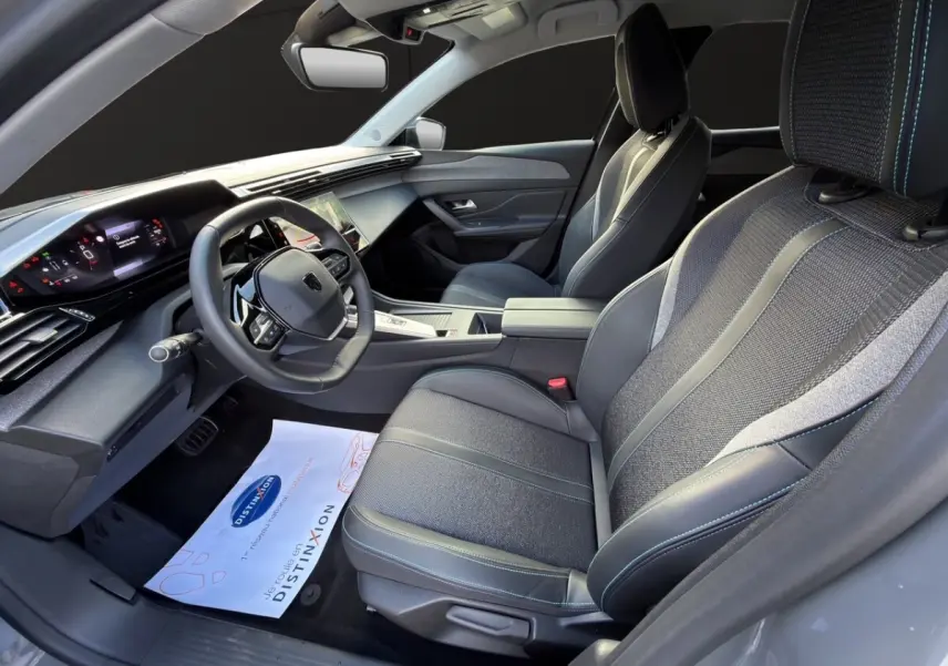 Vue intérieure côté conducteur de la Peugeot 308 Hybrid 2025, sièges tissu et cuir gris avec surpiqûres bleues.