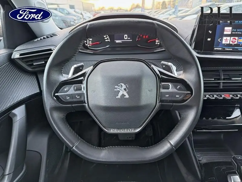 Volant cuir noir avec logo Peugeot au centre, tableau de bord numérique et écran tactile à droite dans un Peugeot 2008 gris Artense 2021.