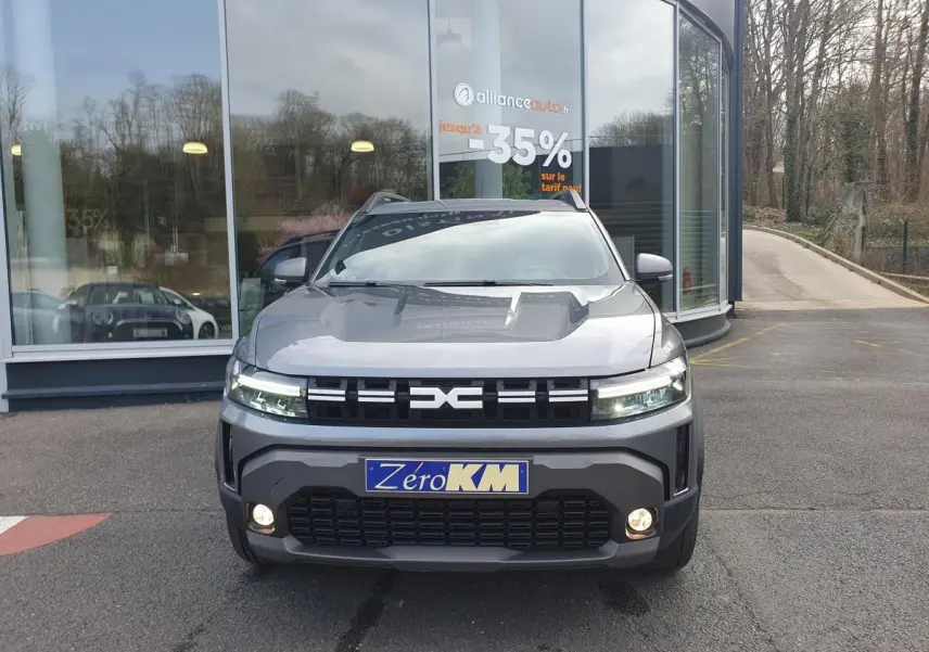 Vue avant d'un Dacia Duster 1.6 Hybrid gris schiste avec calandre noire et feux LED allumés, stationné devant un bâtiment vitré.
