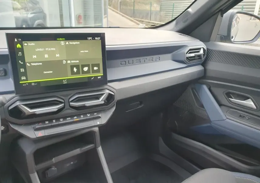Vue intérieure côté passager du tableau de bord gris du Dacia Duster 2026 avec écran tactile et aérateurs hexagonaux.