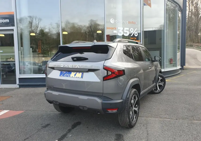 Vue 3/4 arrière droite d’un Dacia Duster gris schiste avec jantes alliage 18” diamantées, stationné devant un showroom.