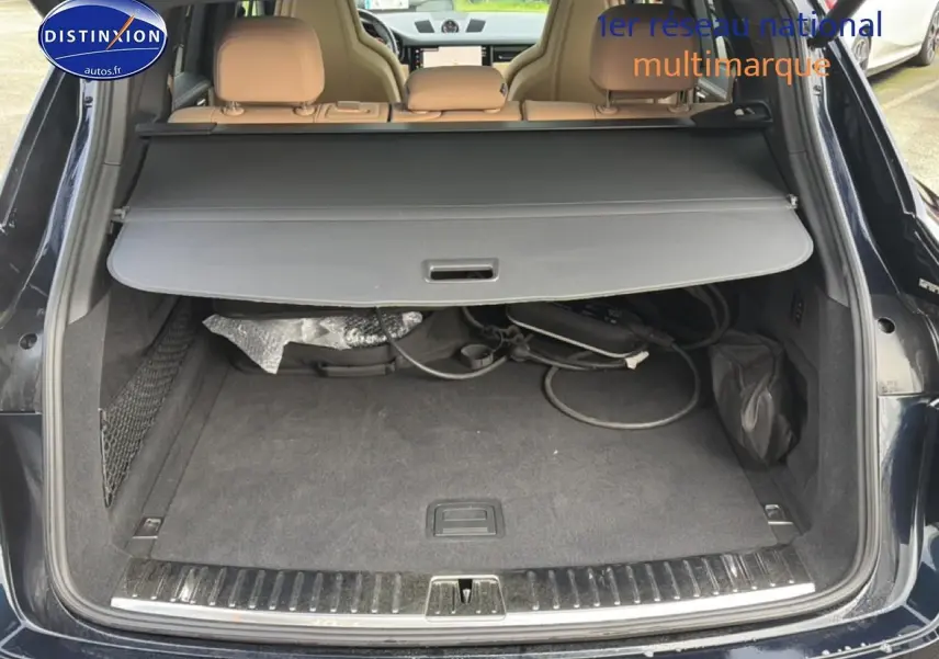 Coffre ouvert d’un Porsche Cayenne bleu, vue arrière, avec cache-bagages et câbles de recharge visibles.