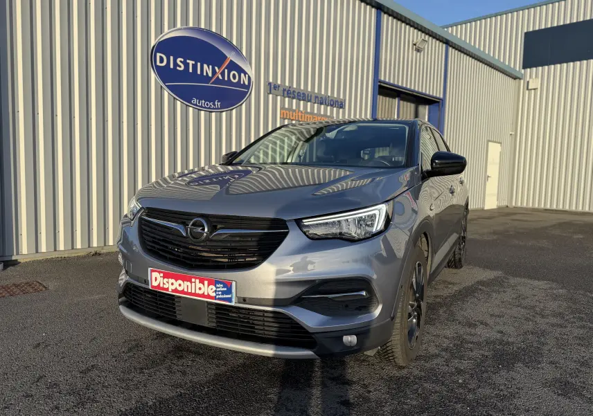 Opel Grandland X gris quartz en 3/4 avant droit, avec calandre noire et jantes foncées, stationné devant un bâtiment industriel.