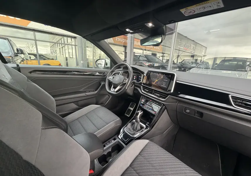 Vue intérieure côté conducteur du Volkswagen T-Roc Cabriolet blanc, mettant en valeur le tableau de bord moderne et l'écran tactile central.