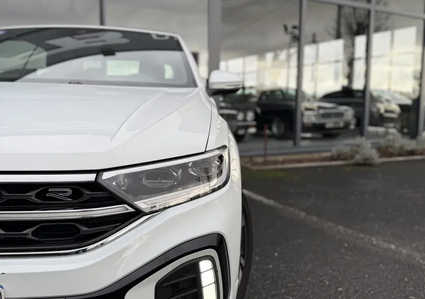 Vue 3/4 avant droit du Volkswagen T-Roc Cabriolet blanc, mettant en valeur le logo R-Line et les feux LED distinctifs.