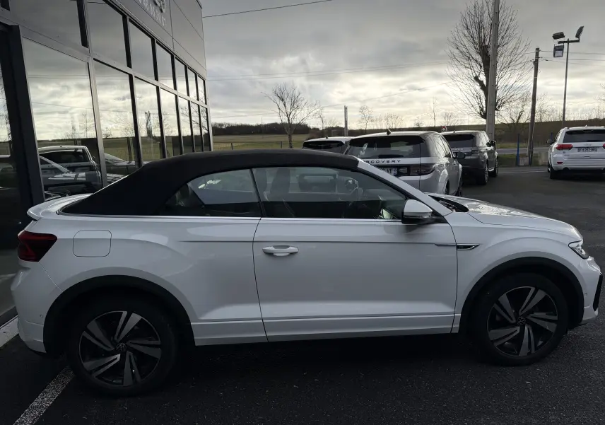 Vue de profil côté gauche d’un Volkswagen T-Roc Cabriolet blanc avec toit noir fermé, version R-Line 2022, stationné devant un bâtiment vitré.