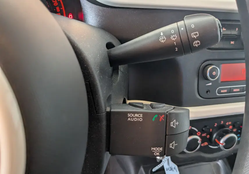 Gros plan sur la commande audio et le commodo d’essuie-glace à gauche du volant dans l’habitacle noir de la Renault Twingo III.