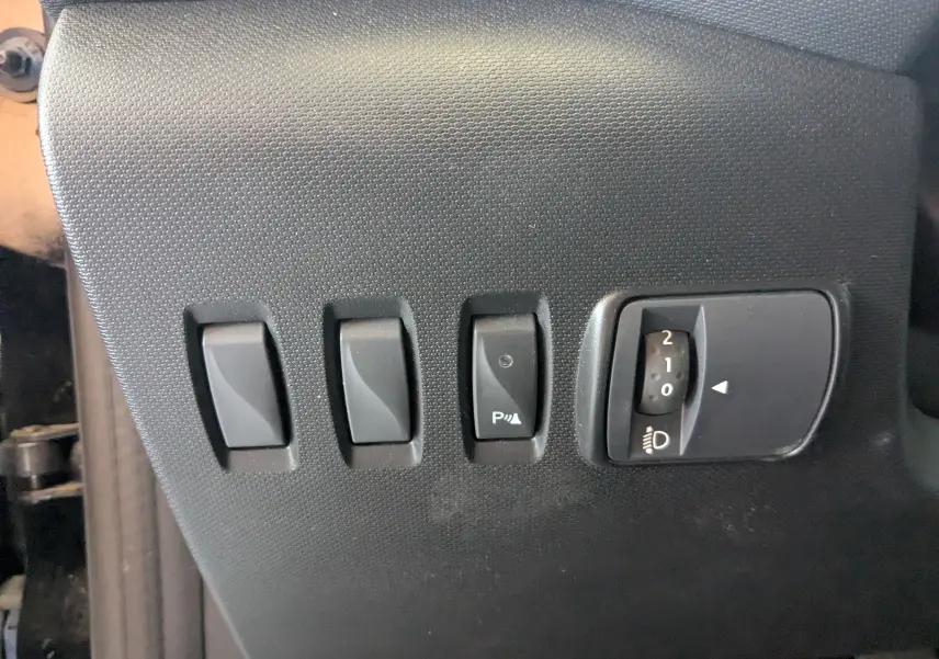 Gros plan sur les commandes de réglage des phares et du système de stationnement d'une Renault Twingo III noire, côté conducteur.