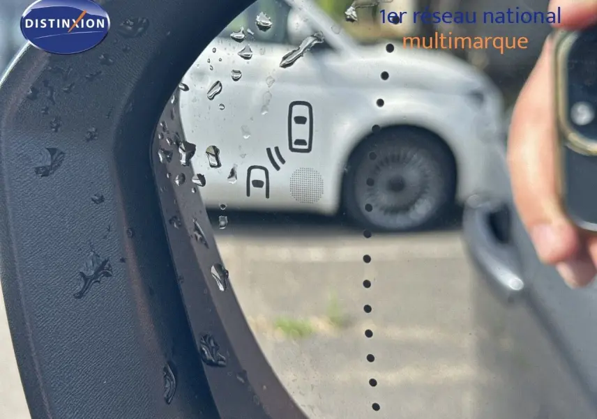 Reflet dans le rétroviseur côté gauche montrant une voiture blanche floue avec gouttes d'eau sur le miroir.