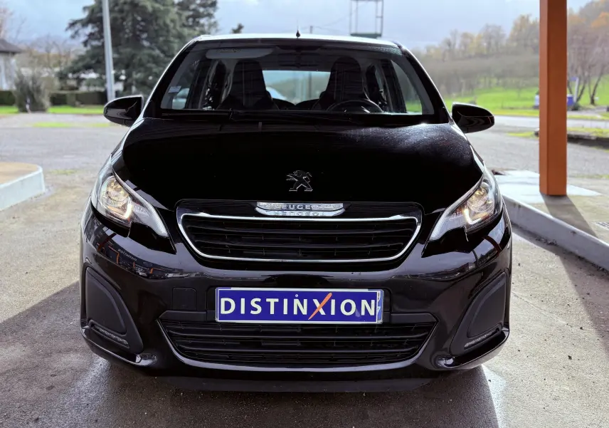 Vue avant d'une Peugeot 108 noire 2018 avec plaque Distinxion et phares allumés sous abri.