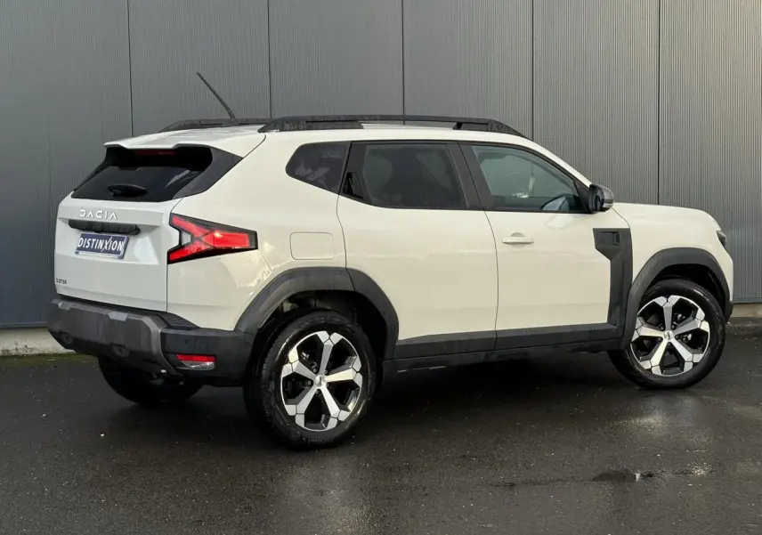 Vue 3/4 arrière droite du Dacia Duster 2024 beige Sandstone avec jantes alliage et barres de toit noires.
