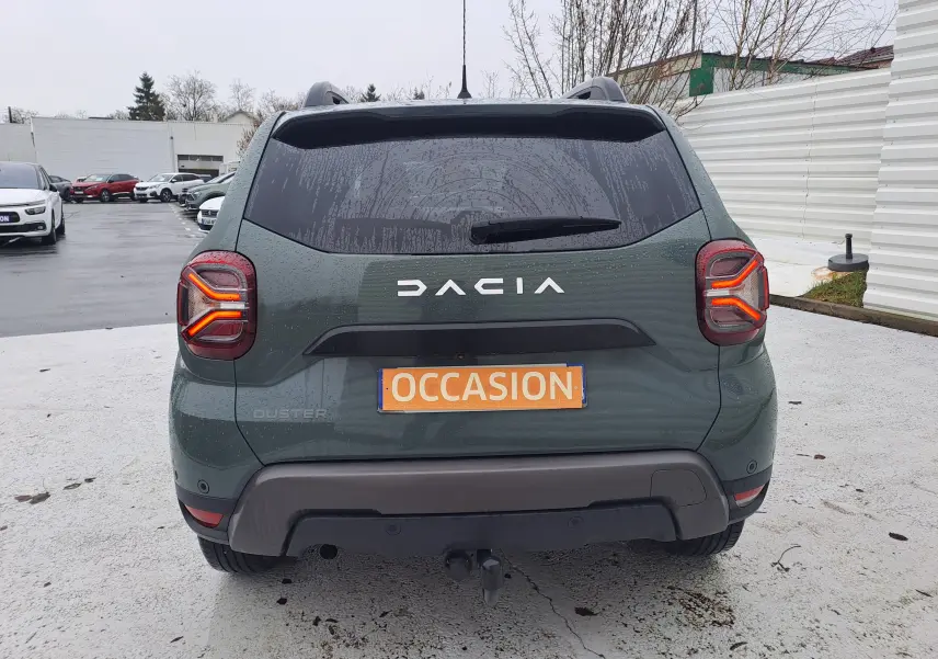 Vue arrière d'un Dacia Duster 2023 kaki Liken avec attelage et feux arrière allumés sur parking humide.