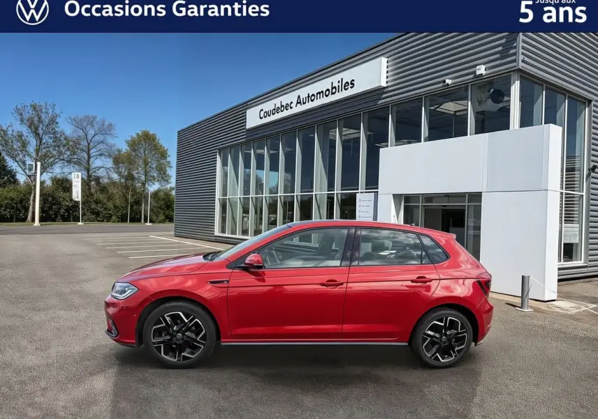 Vue de profil côté gauche d'une Volkswagen Polo rouge métallisée garée devant un bâtiment moderne de concession.