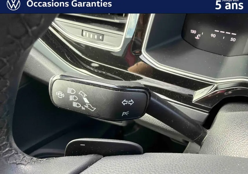 Gros plan sur la commande de clignotants et feux, avec tableau de bord digital partiel d'une Volkswagen Polo rouge.