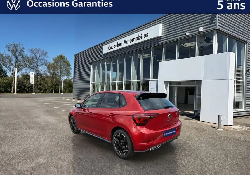Vue 3/4 arrière droite d'une Volkswagen Polo rouge métallisée garée devant un bâtiment moderne de concession.