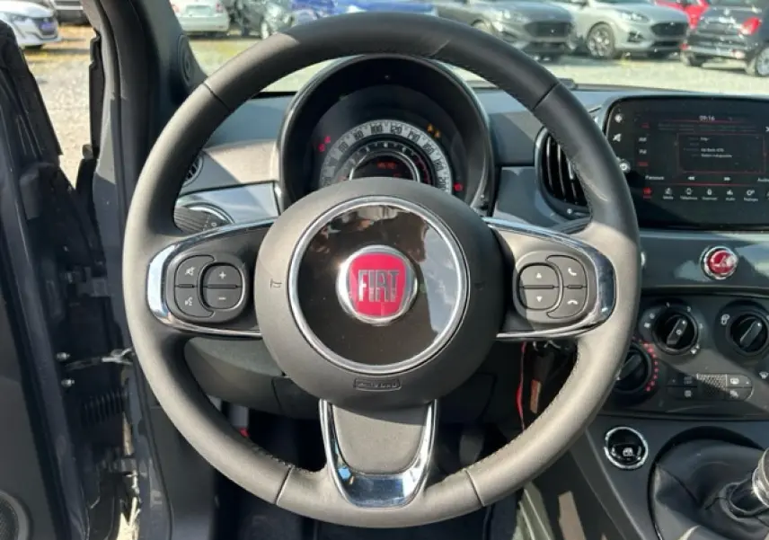Vue intérieure centrée sur le volant noir avec logo FIAT rouge, tableau de bord et console centrale d'une 500C gris/rouge.