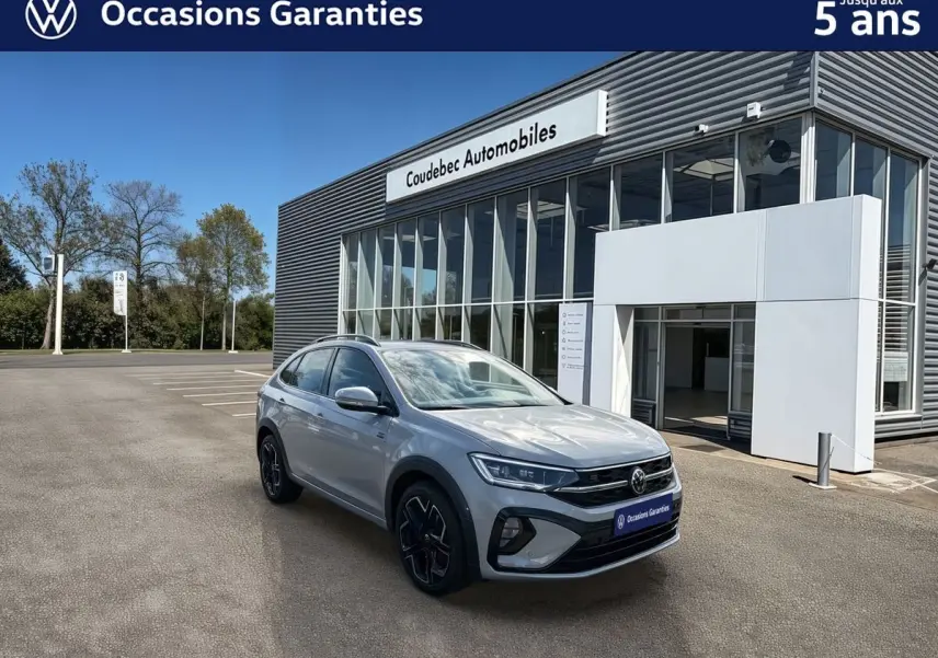 Volkswagen Taigo gris en 3/4 avant droit, avec jantes alliage noires, devant un bâtiment de concession Coudebec Automobiles.