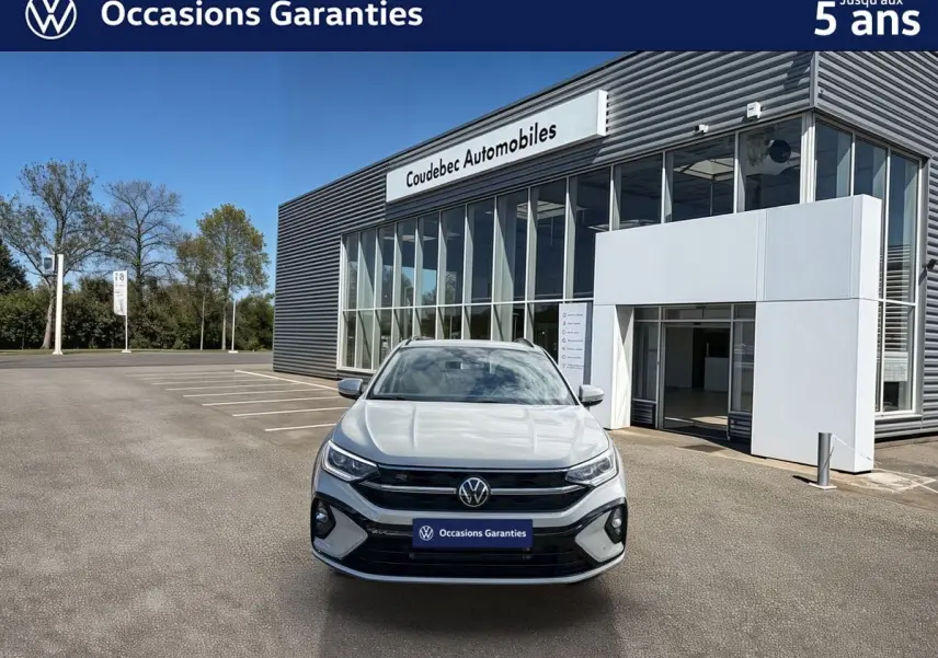 Vue frontale d'un Volkswagen Taigo gris devant un bâtiment moderne de concession automobile par temps clair.