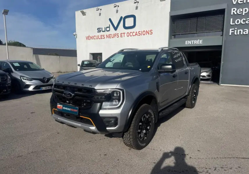 Ford Ranger Wildtrak gris vu en 3/4 avant droit, avec barres de toit et jantes alliage noires visibles.