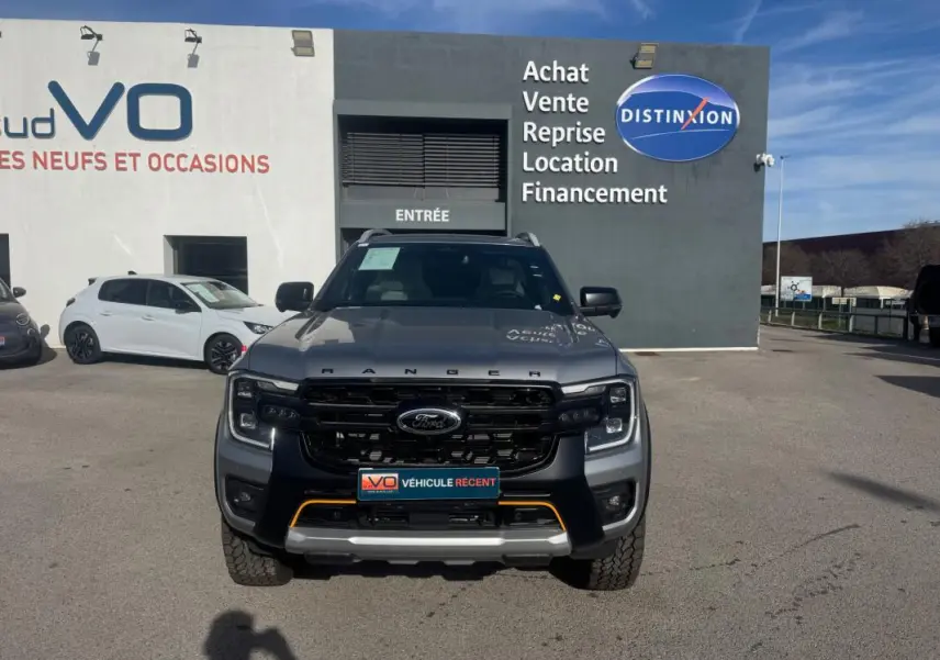Vue avant d'un Ford Ranger gris 2025 Wildtrak X avec calandre noire et barres de toit, stationné devant un concessionnaire.