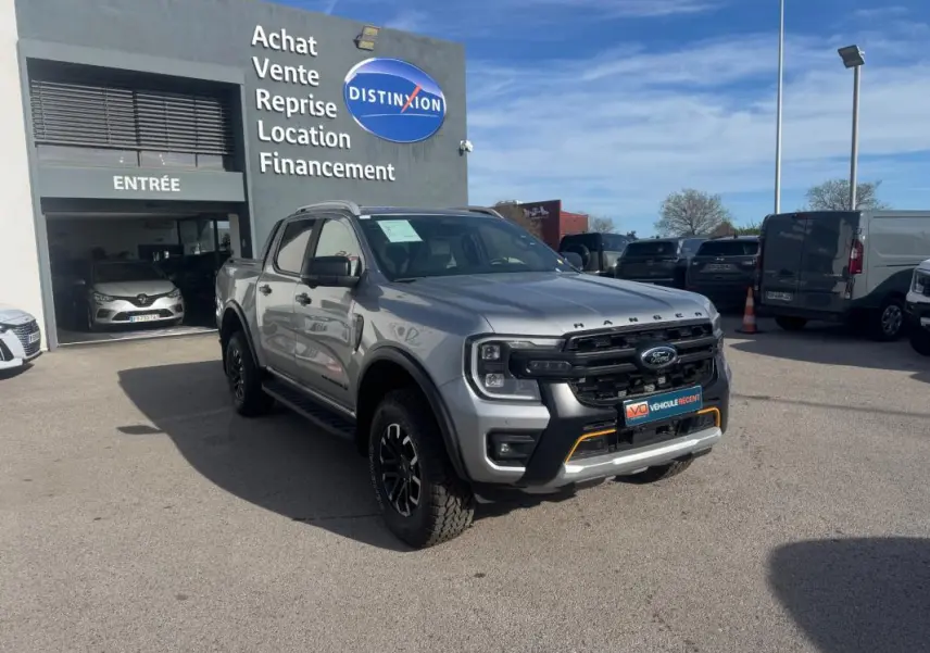 Ford Ranger Wildtrak gris en 3/4 avant droit, avec barres de toit et jantes alliage noires visibles.