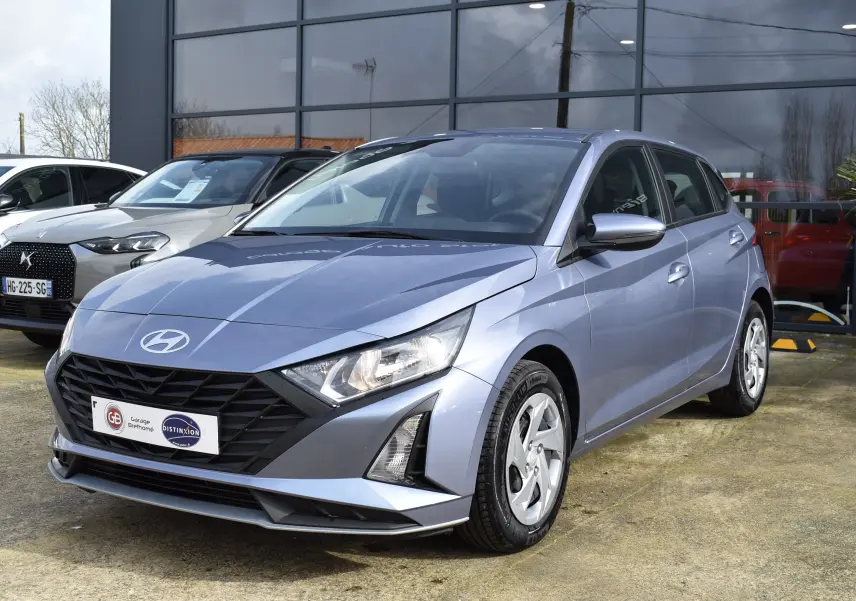 Hyundai i20 bleu clair vue 3/4 avant droit, avec calandre noire et phares anguleux allumés.