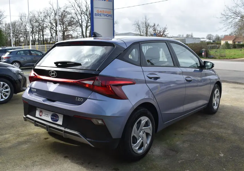 Vue 3/4 arrière droite d'une Hyundai i20 bleu clair 2025 avec spoiler de toit et feux arrière distinctifs en forme de flèche.