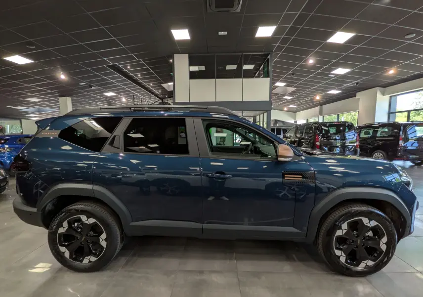 Vue de profil droit d'un Dacia Bigster bleu indigo avec jantes noires bicolores et détails noirs, en showroom.