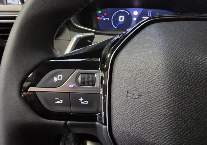 Gros plan sur les commandes gauche du volant cuir noir de la Peugeot 208 hybride 2025 avec tableau de bord numérique.