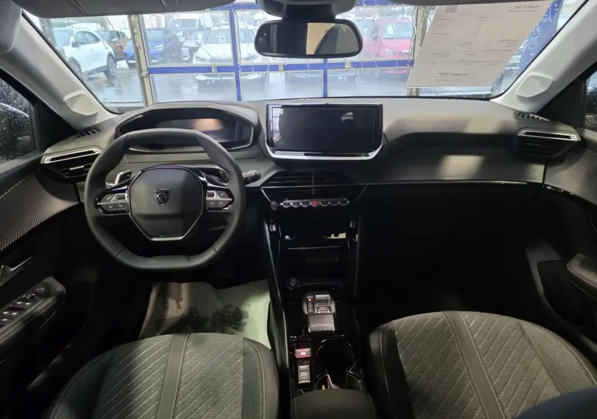 Vue intérieure avant de la Peugeot 208 1.2i Hybrid 2025, tableau de bord noir avec volant compact et écran tactile central.
