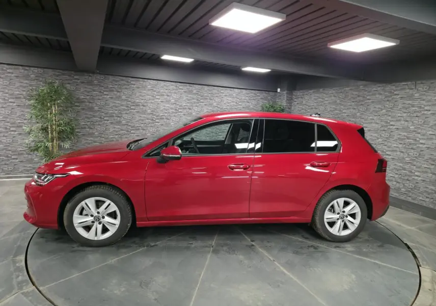 Profil droit d'une Volkswagen Golf VIII rouge métallisée, avec vitres arrière surteintées dans un showroom.