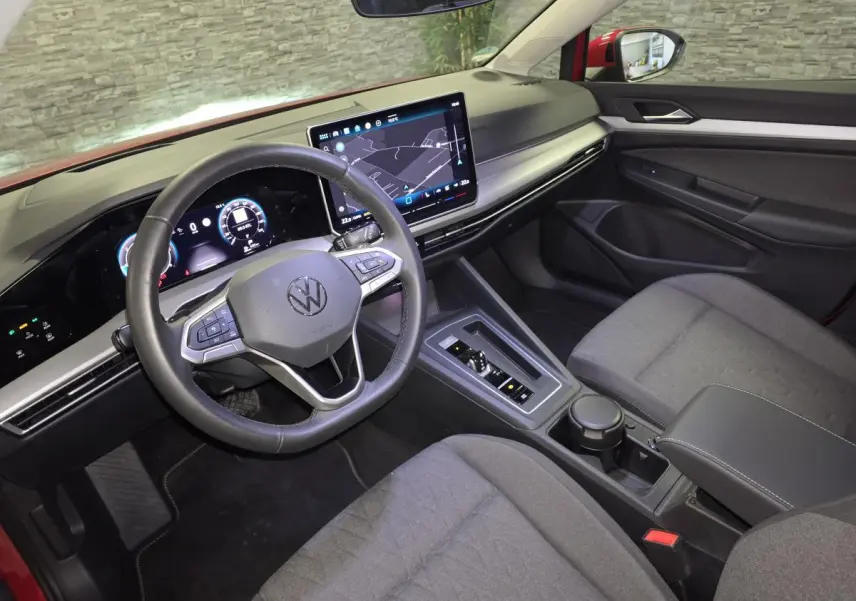 Vue intérieure côté conducteur de la Volkswagen Golf VIII rouge, avec tableau de bord digital et écran tactile central.