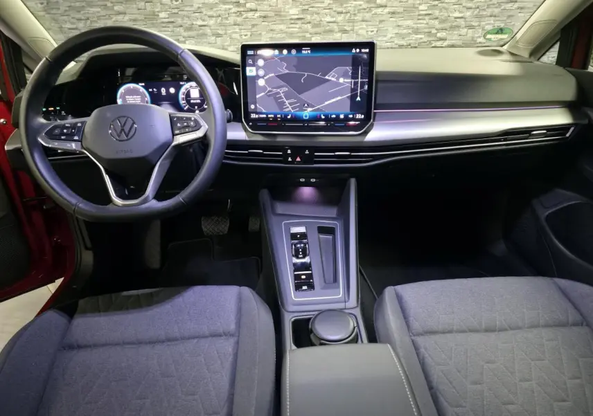 Vue intérieure avant de la Volkswagen Golf VIII rouge, tableau de bord moderne avec écran tactile central et volant multifonction.