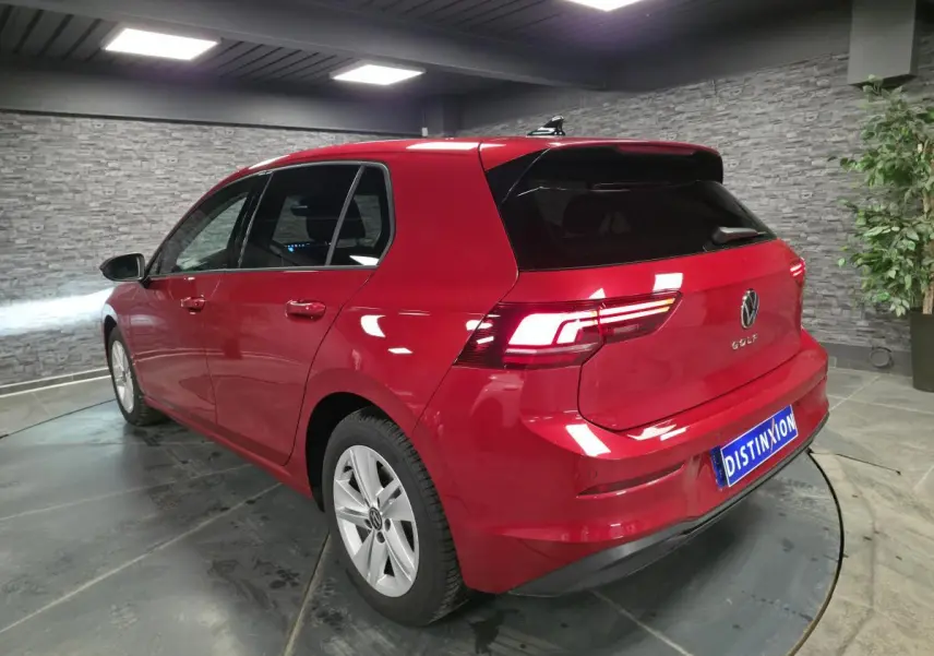 Vue 3/4 arrière droite d'une Volkswagen Golf VIII rouge métallisée dans un showroom avec vitres arrière surteintées.
