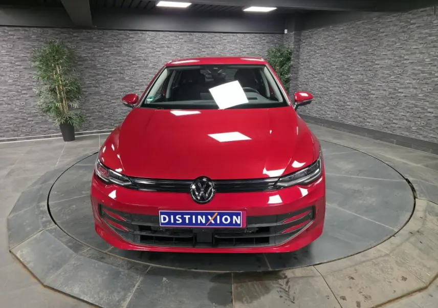 Volkswagen Golf VIII rouge métallisé vue de face dans un showroom avec éclairage intérieur et mur en pierre grise.